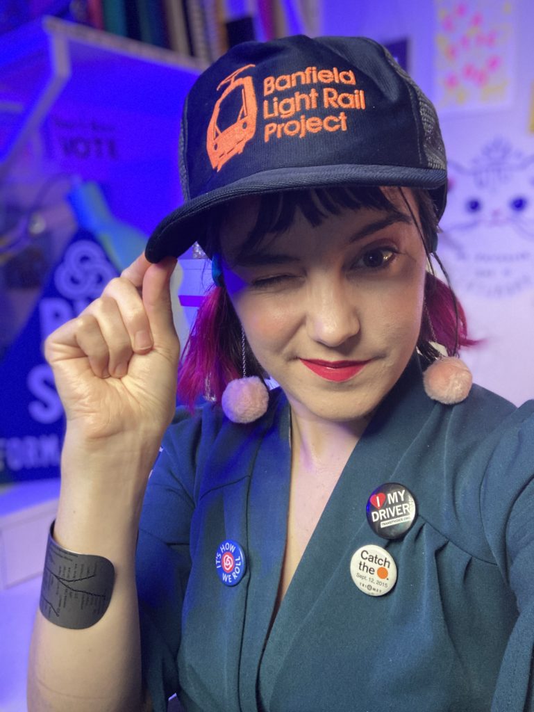 Lillian looking into the camera winking while wearing a blue hat that says "Banfield light rail project" and several transit buttons