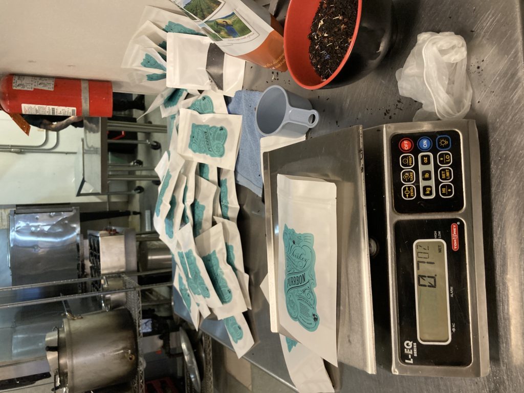 A commercial kitchen counter showing a bowl of tea, a commerical food scale and dozens of bags of tea being packed.