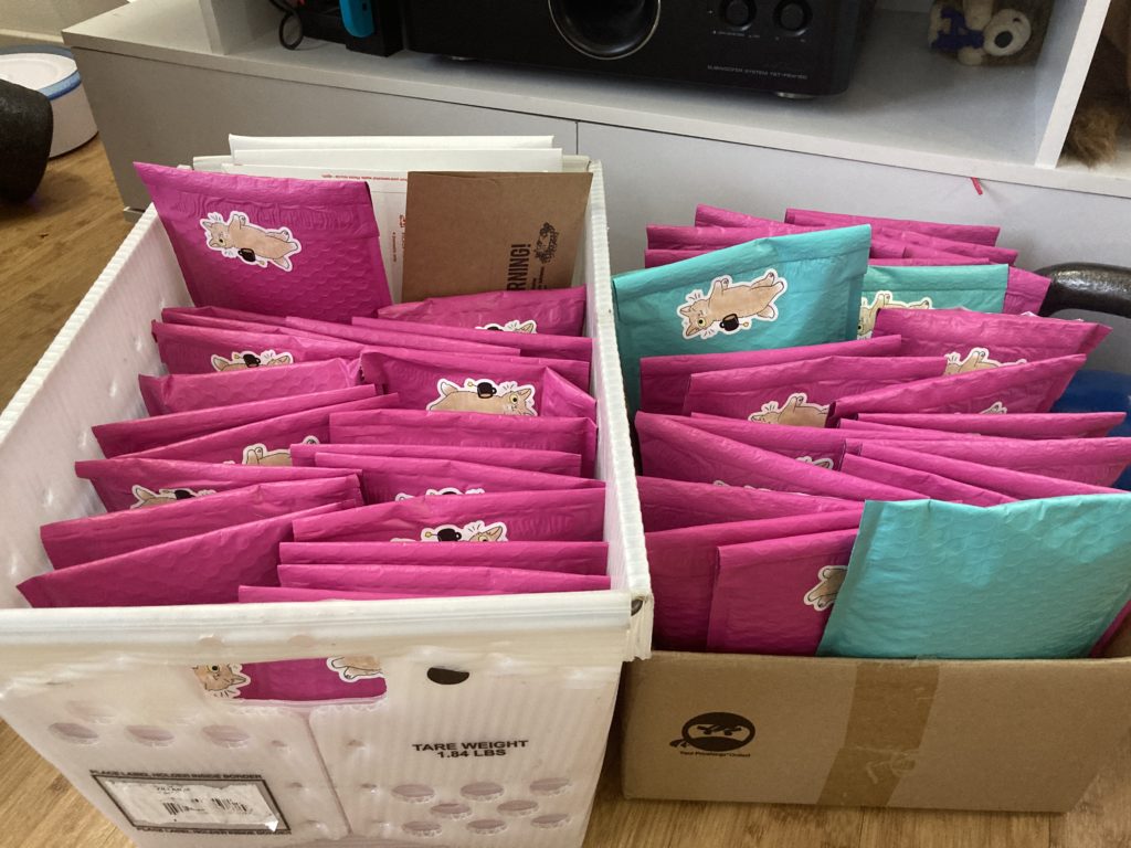 Two boxes full of pink bubble mailers with stickers of cats drinking tea on them.