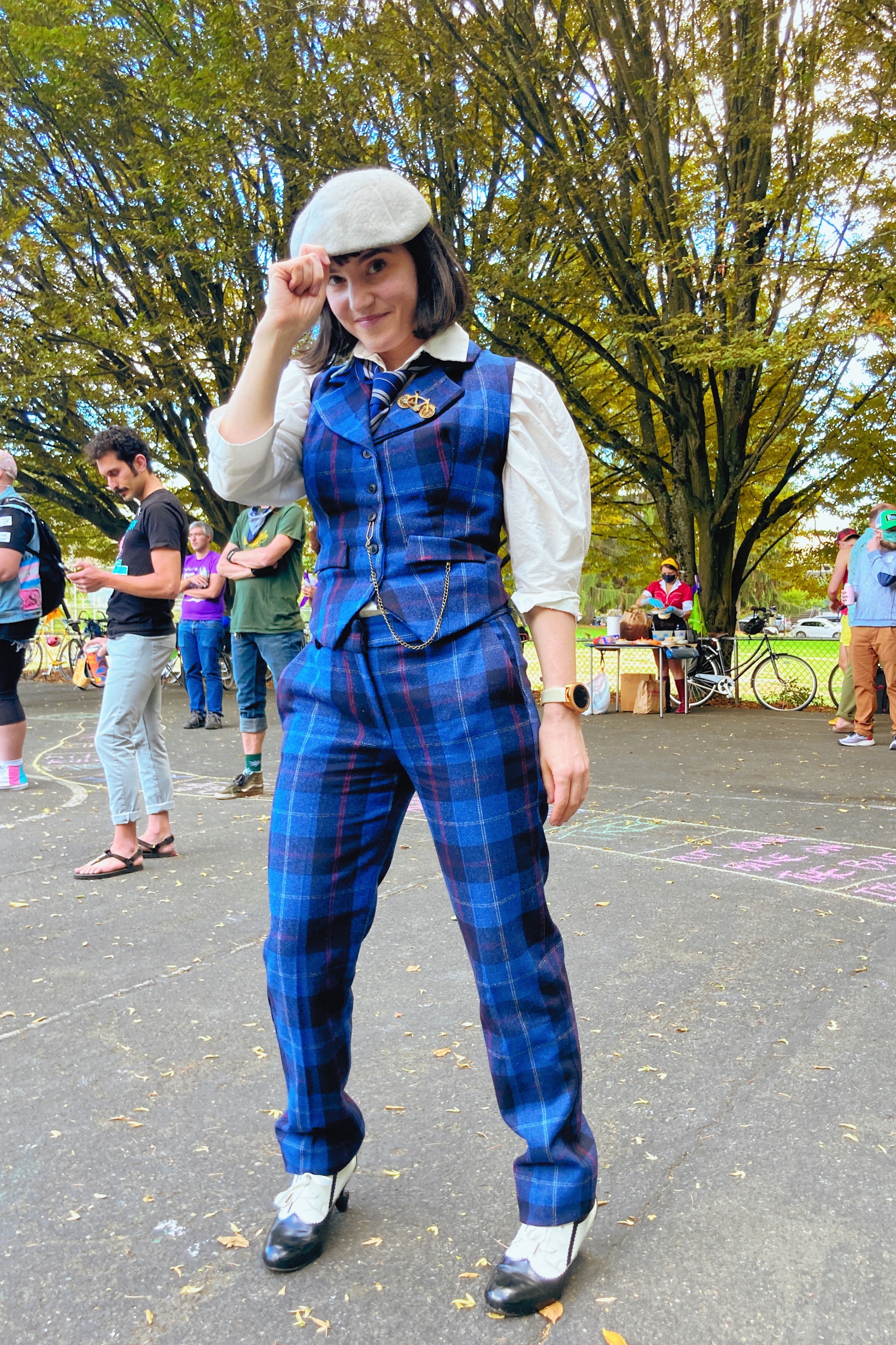 Me in a blue plaid waistcoat and blue plaid pants, black and white oxford shoes and gray flatcap. Standing in front of a group of people and trees on a sunny day